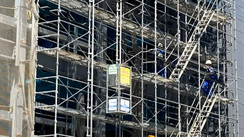 Workers assembling scaffolding at a Japanese construction site using tube and clamp scaffolding with temporary access ladders.