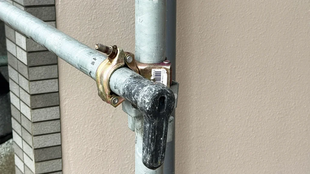 Close-up scaffold connection detail showing metal tubes joined by a clamp fitting beside an exterior building wall.
