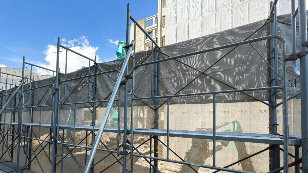 Real scaffold showing repeated bays, standards, working platforms, diagonal bracing and protective mesh on a construction site.