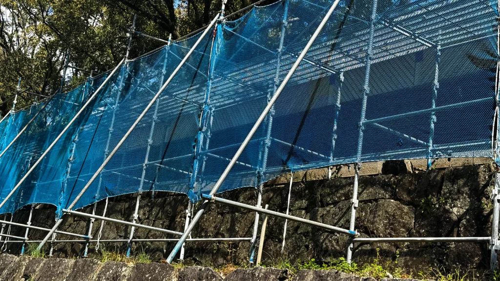 Scaffold with diagonal bracing and blue mesh installed along irregular sloping ground